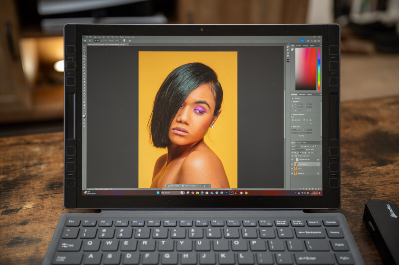 A laptop displaying an image-editing software on its screen. The software shows a portrait of a person with bold makeup, including purple eyeshadow and pink lipstick, against a yellow background. A black keyboard is visible in the foreground.