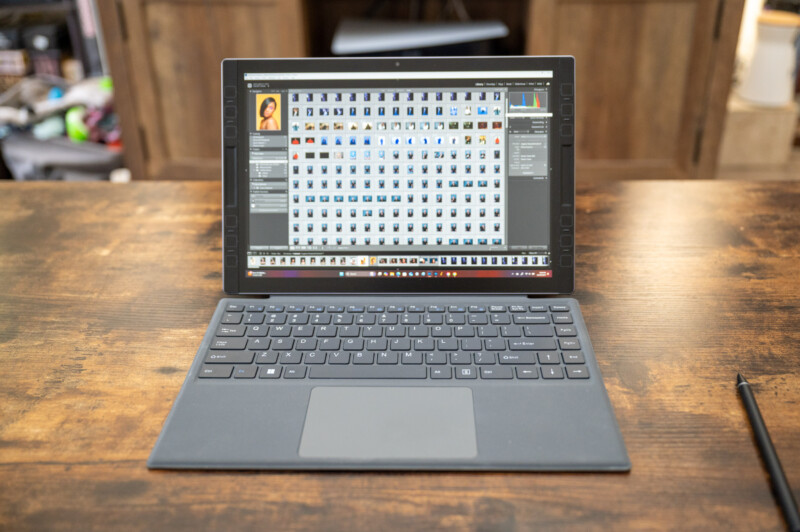 A laptop sits on a wooden table, displaying photo editing software with a grid of images on its screen. A stylus lies nearby, and wooden furniture is visible in the background.