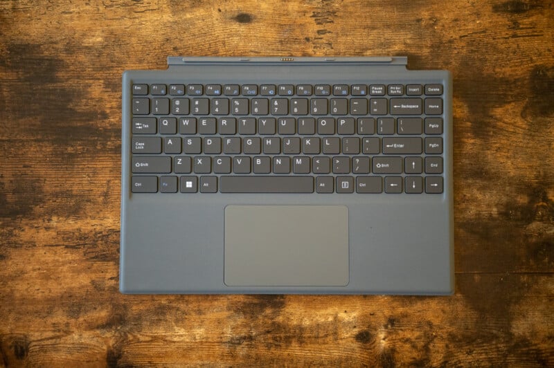 A detachable black keyboard with a touchpad is placed on a rustic wooden surface. The keyboard layout includes standard keys and function keys, with a sleek and compact design.