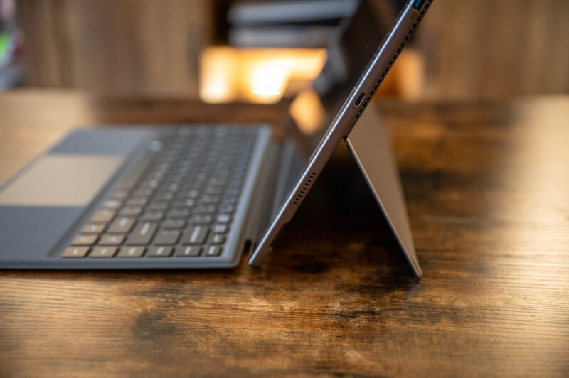 A close-up side view of a tablet with a keyboard accessory, set up on a wooden table. The tablet is propped up using its built-in kickstand, with soft, warm lighting in the background.