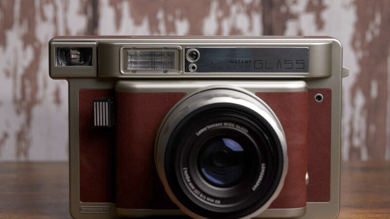 Close-up of a vintage-style camera with a large lens, featuring a brown leather body and silver accents. The camera is set against a blurred wooden background, creating a rustic aesthetic.