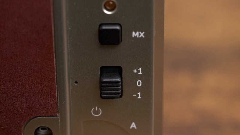 Close-up of a device panel with a black switch labeled MX and a sliding switch with settings +1, 0, and -1. Below is a power button symbol with a light gray letter A on a dark background.