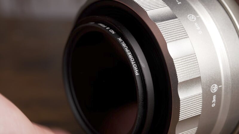 Close-up of a camera lens showing a PHOTOREPUBLIK ND filter attached. The lens barrel is silver with focus distance markings, and the focus is on the filter text. The background is blurred.