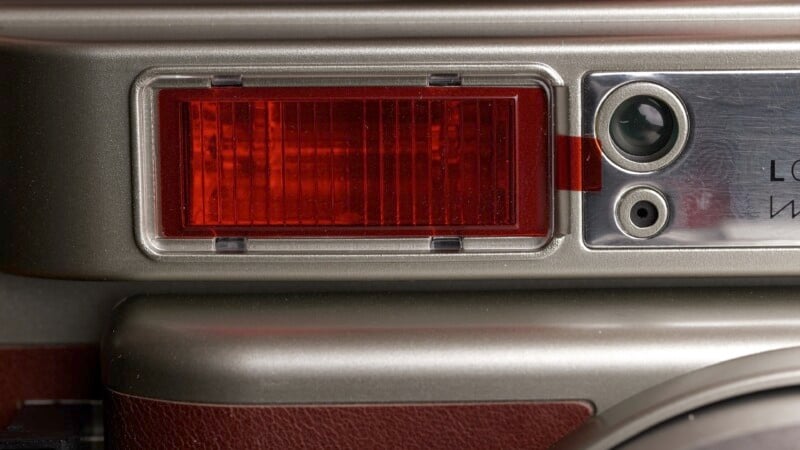 Close-up of a vintage camera featuring a red rectangle lens, a round viewfinder, and metallic frame with a leather-texture accent. The design is classic with a combination of silver and maroon colors.