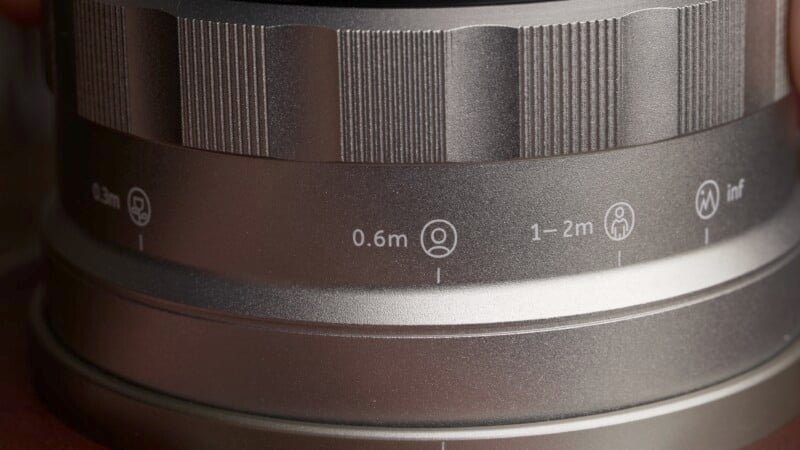 Close-up of a camera lens with focus distance markings. The markings range from 0.5m to infinity, including icons for portrait and landscape distances. The lens surface is metallic with textured grip areas.