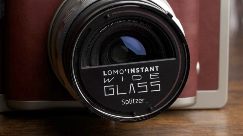 Close-up of a camera lens with the text "LOMO'INSTANT WIDE GLASS Splitzer" printed on it. The camera body is partially visible in the background, featuring a brown and beige color scheme.