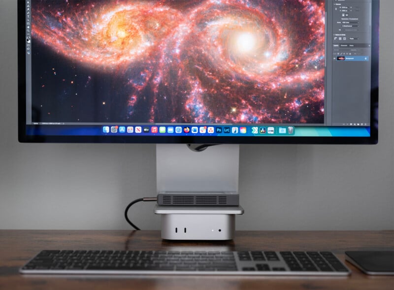 A computer monitor displaying a vibrant galaxy image is on a desk. Below the monitor, a small computer tower and a wireless keyboard are visible. The galaxy image features bright, swirling star patterns in red, orange, and white hues.