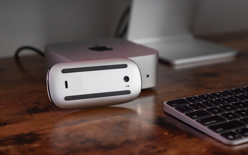 A computer mouse is placed upside down on a wooden desk next to a keyboard. In the background, a monitor is partially visible, connected to a compact computer tower. The setup has a sleek, modern design.