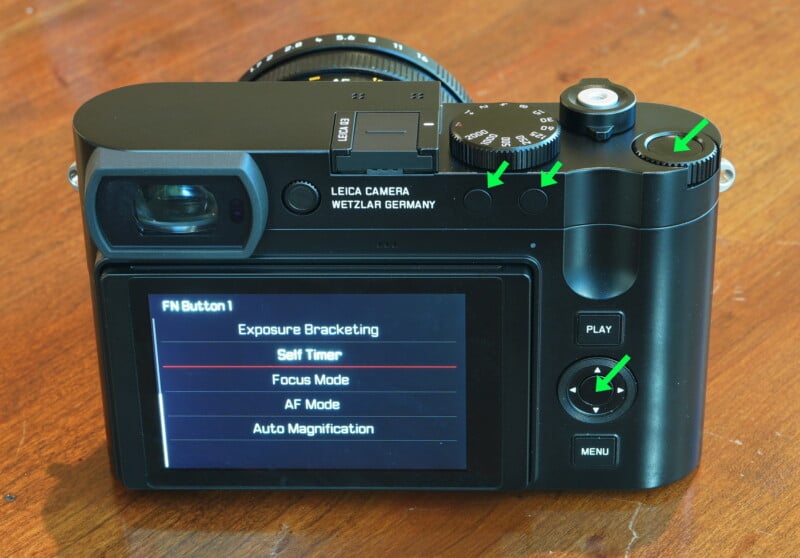 Back view of a Leica camera showing an LCD screen with menu options like "Self Timer" and "Focus Mode." Green arrows point to buttons on the top and back of the camera. The surface is a wooden table.