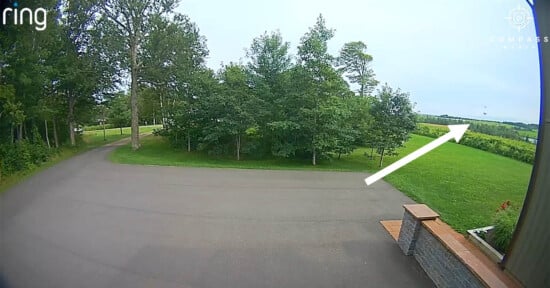 A wide driveway curves toward lush green trees under a cloudy sky. A white arrow overlays the image, pointing towards an open grassy area to the right, bordered by more trees.