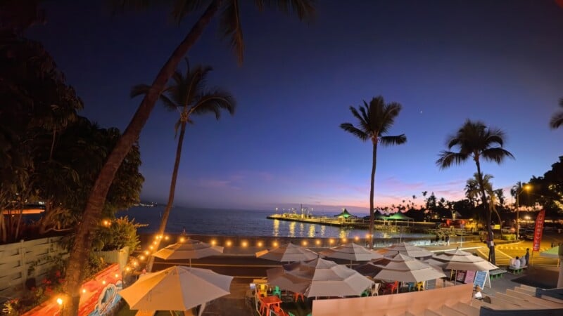 A tropical beach scene at dusk with vibrant pink and blue skies. Palm trees silhouetted against the horizon, beach umbrellas in the foreground, and string lights illuminating the area. The ocean is calm, with a distant pier visible.