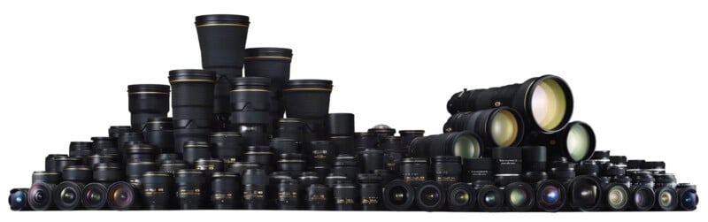 A large assortment of camera lenses of various sizes and types are arranged in rows, forming a pyramid-like shape against a white background. The lenses range from wide-angle to telephoto, showcasing their reflective glass elements.