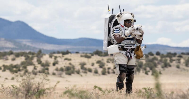 An astronaut in a spacesuit walks on a rocky, arid landscape with mountains in the background during daylight. The person is holding a device, possibly conducting experiments or navigation. Sparse vegetation is visible in the foreground.