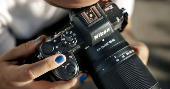 A person holds a Nikon camera, adjusting the settings on the top dial. The person's nails are painted blue. The lens is large and the camera is in use, suggesting an outdoor or photography setup.