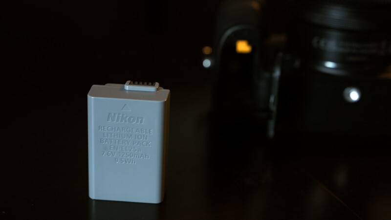 Close-up of a Nikon rechargeable lithium-ion battery pack (EN-EL23) standing upright. The background is dimly lit, featuring a blurred camera on the right. The battery text indicates 7.6V, 1250mAh, and 9.5Wh.