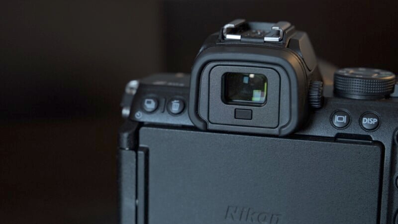 Close-up of a black Nikon camera focused on the electronic viewfinder, featuring a textured surface and buttons on the side. The LCD screen is slightly tilted, and the brand name is partially visible. Dark background.