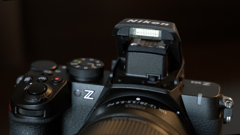 A close-up of a Nikon Z series camera shows the camera's top controls and an open flash unit. The lens is partially visible, and the focus is on the camera's buttons and flash mechanism. The background is blurred and dark.