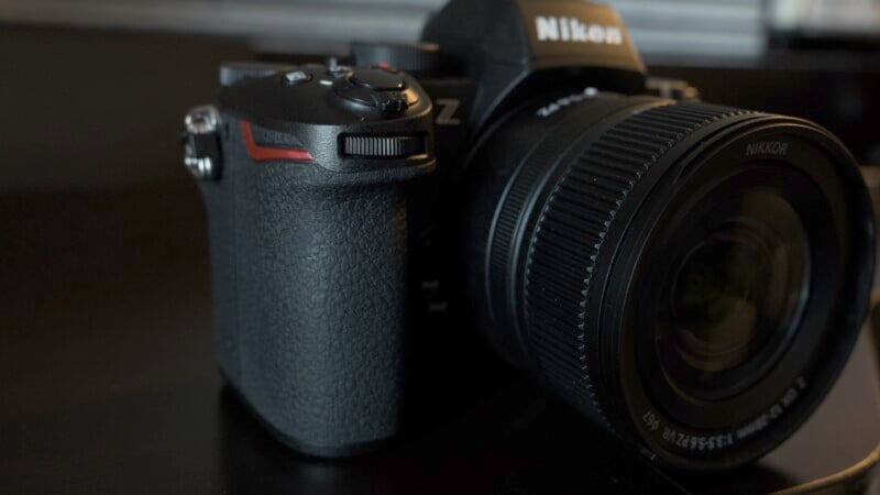 Close-up of a Nikon camera with a textured grip and visible lens, positioned on a dark surface. The camera is partially illuminated, highlighting its brand and sleek design.