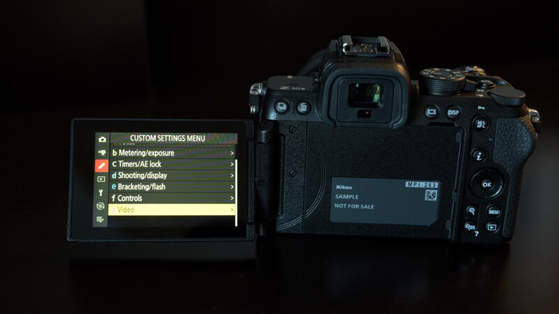 A digital camera with a flip-out screen showing the custom settings menu. Options include metering exposure, timers, shooting display, bracketing/flash, controls, and video. The screen displays a sample message and a model identifier.