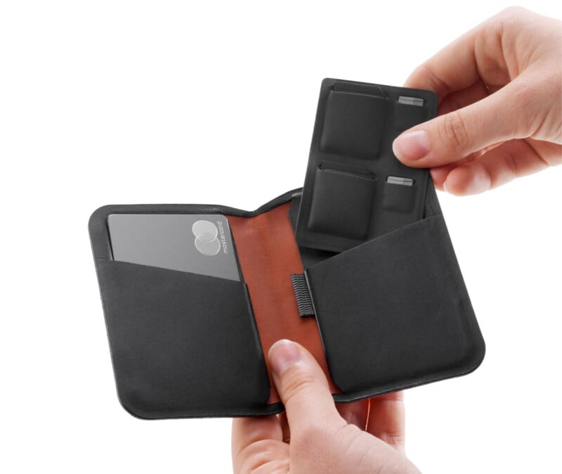 A person holds a black wallet open, displaying compartments. A credit card is in one pocket, and the other contains slots for items. The interior is brown with a fabric band in the center. The person's fingers are visible holding the wallet.