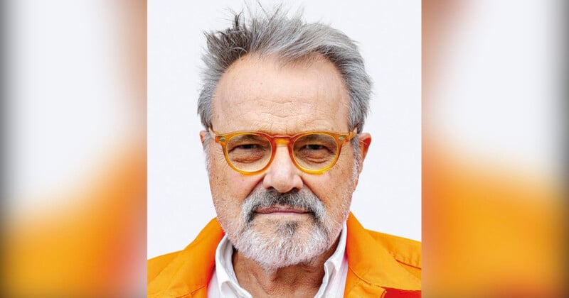 A man with gray hair and a beard wearing orange-rimmed glasses and a bright orange jacket looks directly at the camera against a plain background.