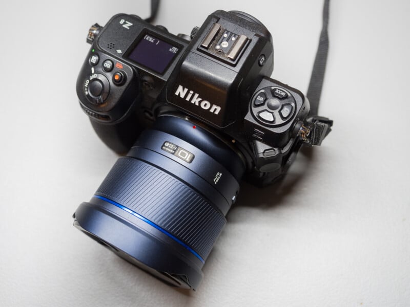 A Nikon Z camera with a mounted lens on a light-colored surface. The camera has several buttons and a small screen on top, along with a visible Nikon logo. Its strap is partially visible.