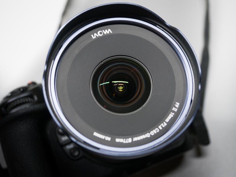 Close-up of a camera lens with a Laowa brand label. The lens is on a camera body, and the focus ring details are visible, including aperture and focal length markings. The image emphasizes the circular shape and intricate design of the lens.