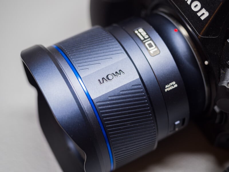 Close-up of a dark blue Laowa camera lens attached to a Nikon camera. The lens has a textured grip and "Auto Focus" text on its side. The lens reflects light, highlighting its sleek design.