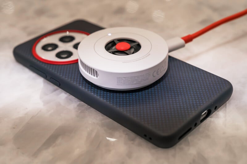 A smartphone with a textured dark case on a marble surface is wirelessly charging using a white circular charger with a red button and cord.