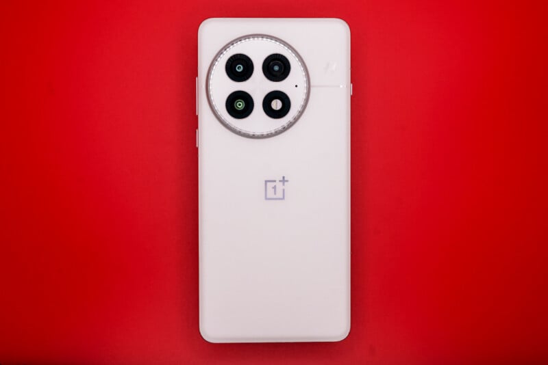 A white smartphone with a circular camera module featuring three lenses and a flash is placed against a red background. The back of the phone displays a logo in the center.