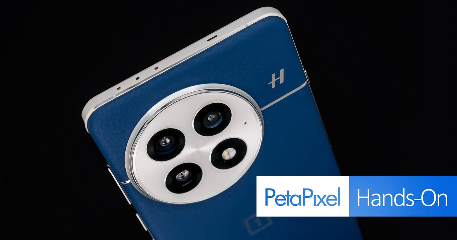 Close-up of a smartphone's rear camera module with three lenses on a sleek, dark blue back. The phone has a metallic frame and a small logo. Text overlay says "PetaPixel Hands-On.