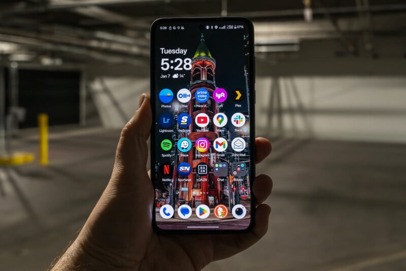 A person is holding a smartphone displaying a colorful wallpaper of a building at night. The screen shows various app icons, including Gmail, WhatsApp, and Spotify. The background is a dimly lit parking garage.