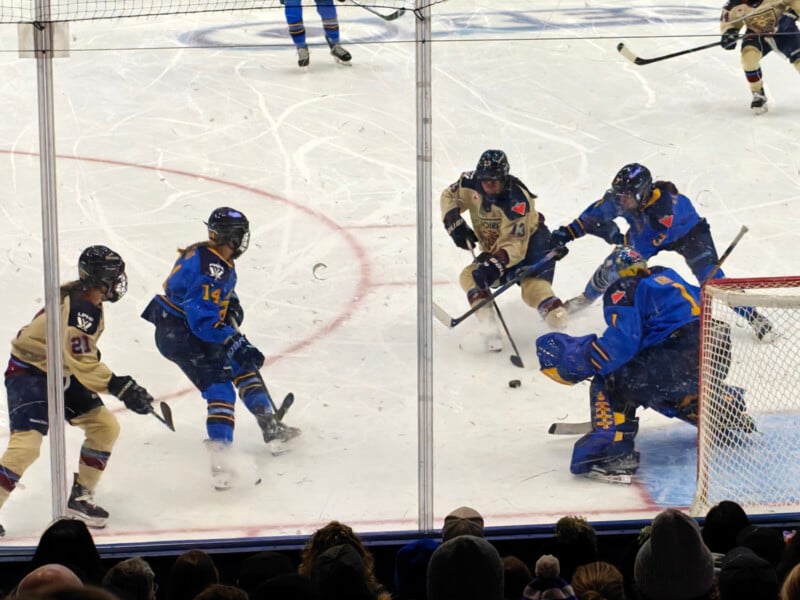 A women's ice hockey game in progress. Players in blue are defending their goal against players in beige. Several players are attempting to gain control of the puck near the goal crease, with spectators visible in the foreground.