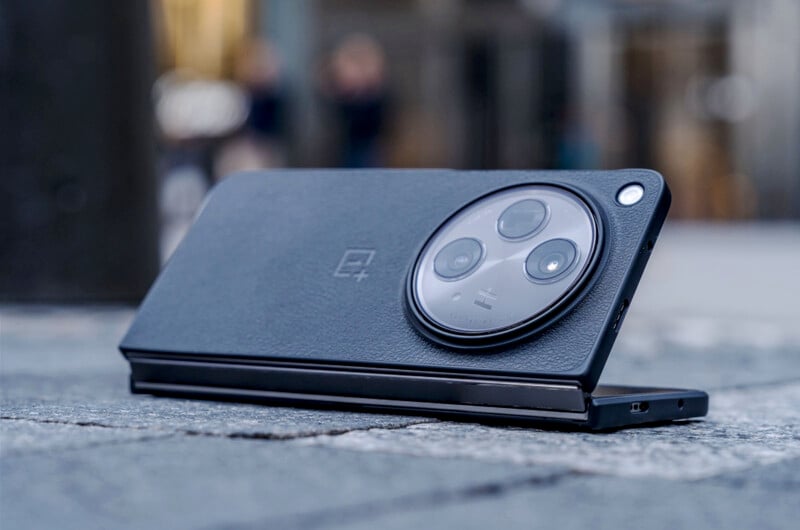 A sleek, black smartphone with a large circular camera module is placed on a cobblestone surface. The phone is partly open, showcasing its foldable design, with the brand's logo subtly visible on the back. Blurred urban background.