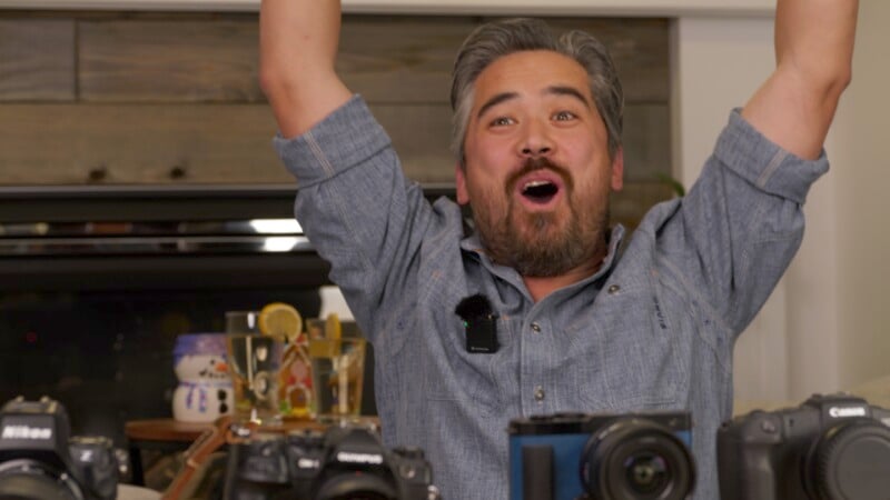 A man with gray hair and a beard looks excited, raising both arms in the air. He is surrounded by various cameras on a table. The background shows shelves with decorative items.