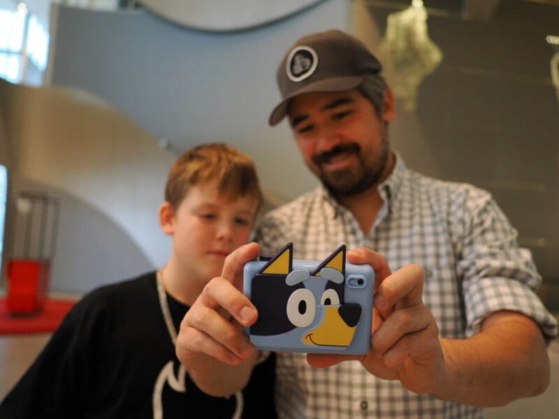 A man and a child take a selfie with a smartphone featuring a cartoon dog case. They are indoors, and both are smiling slightly. The background includes modern architecture elements.