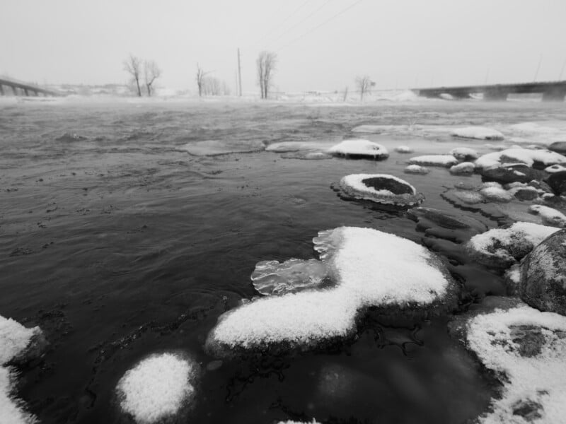 A black-and-white image of a snowy riverbank with rocks partially covered in snow. The river flows gently, and bare trees can be seen in the background against an overcast sky.