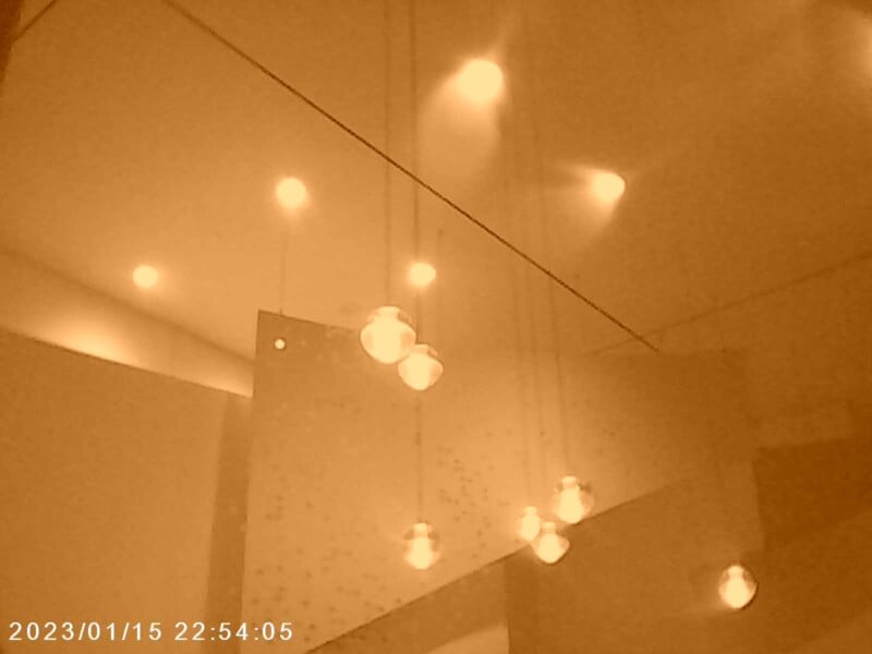 A warm-toned image of a ceiling featuring hanging spherical lights, casting a soft glow. The minimalist decor includes angular lines and recessed lighting. Date and time stamp at the bottom left reads 2023/01/15 22:54:05.