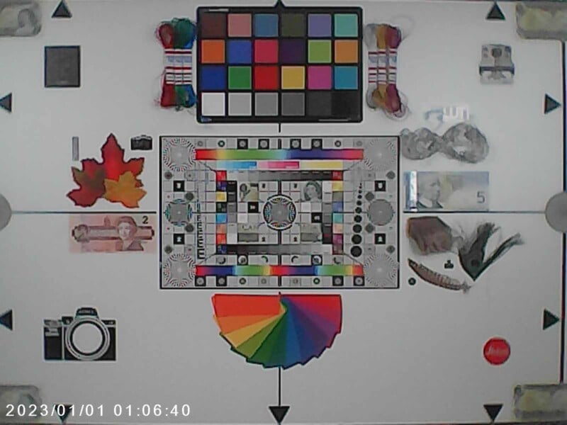 A colorful test chart with various color blocks, grayscale bars, and reference objects such as currency notes, leaves, and feathers. A rainbow color wheel and camera icons are also present. The date "2023/01/01" is on the bottom left corner.