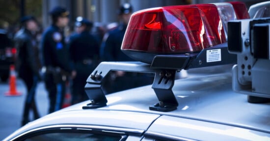 Close-up of a police car's red and blue flashing lights on a city street, with blurred police officers in the background. The scene is set during the day, with buildings visible in the distance.