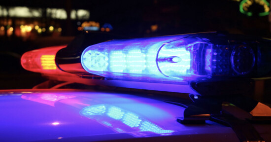 Close-up of a police car light bar at night, with blue and red lights illuminated, reflecting on the vehicle's surface. Blurred lights are visible in the background.