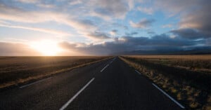 Straight road extending into the horizon, flanked by open fields under a dramatic sky. The sun is setting or rising on the left, casting a warm glow across the landscape and illuminating the clouds above.