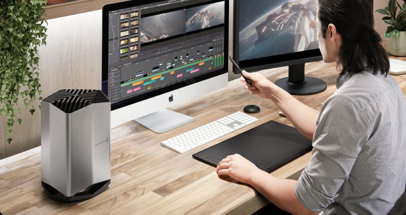 A person with long hair sits at a wooden desk working on video editing software on two monitors connected to a desktop computer. A sleek, silver external device and a pen tablet are also on the desk. Green plants hang in the background.