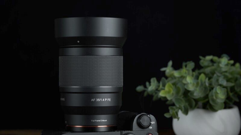 A black camera lens labeled "AF 35/1.4 FE" mounted on a camera against a dark background. A small potted plant with green leaves is blurred in the background, adding a touch of color.