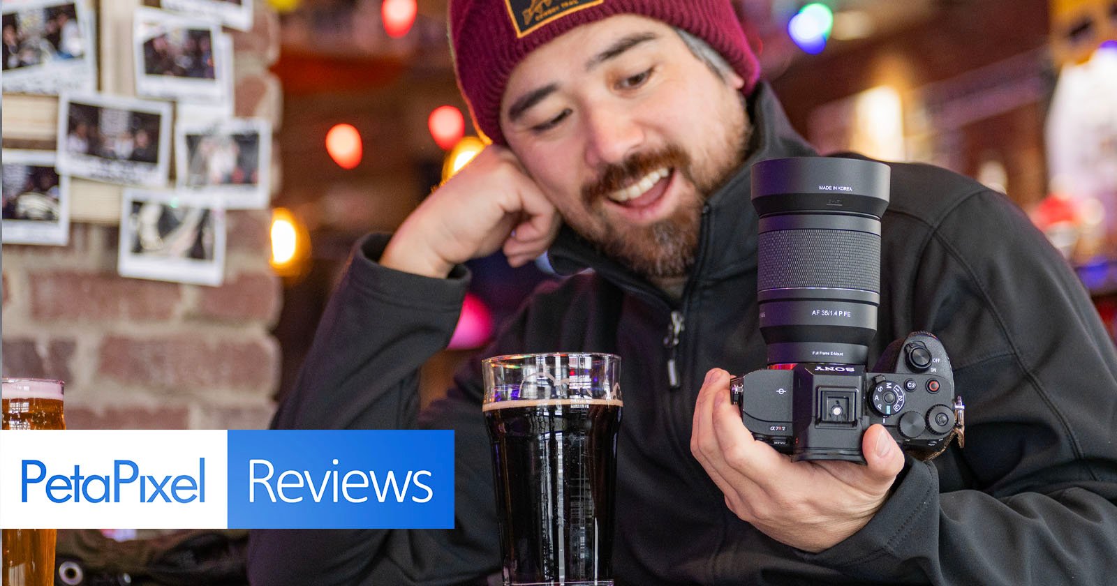 A smiling person in a beanie and black jacket holds a camera in one hand, while sitting at a table with a glass of dark beer. The background features a brick wall with photos and warm lighting. Text: "PetaPixel Reviews.