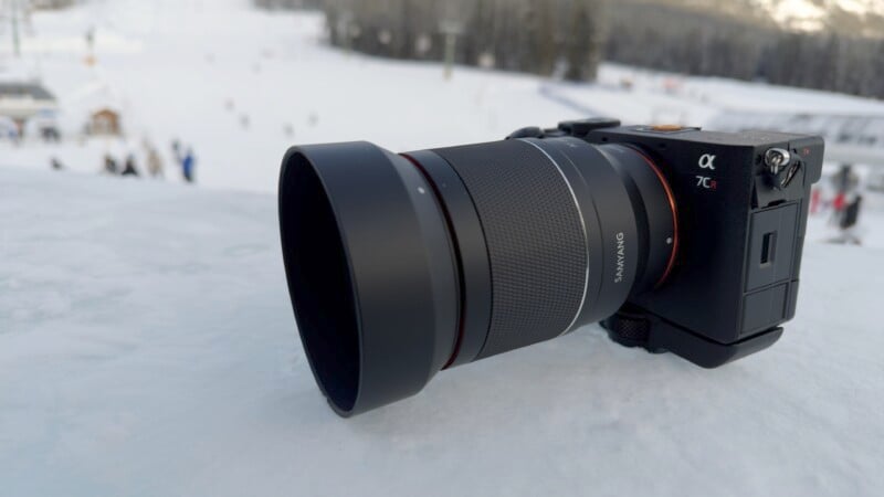 A digital camera with a large lens rests on a snowy surface. The camera and lens have visible branding, and the background shows a snowy landscape with blurry figures and trees, suggesting a winter scene.