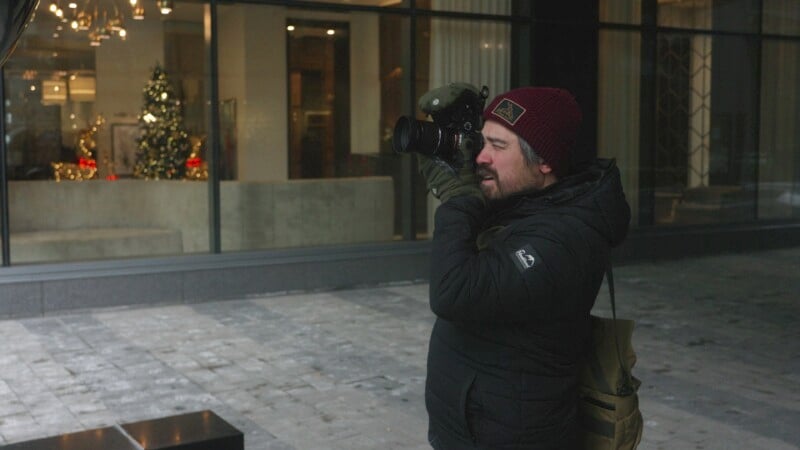 A person in a winter jacket and red beanie takes photos with a camera on a city street. In the background, a window display features a decorated Christmas tree and festive lights.