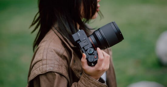 A person with long hair holds a large camera in their right hand. They are wearing a brown jacket and standing outside on a grassy area. The image is focused on the camera, highlighting its detailed features.