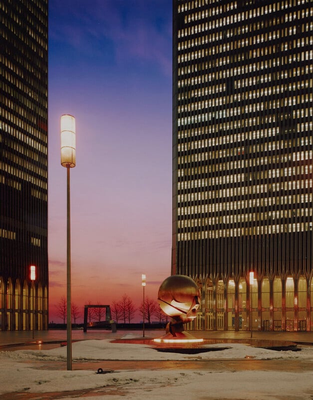 A twilight scene featuring illuminated tall buildings with lit windows. Between the buildings is a bronze spherical sculpture and a streetlamp. The sky showcases a gradient from deep purple to soft orange. The ground appears to be covered with light snow.