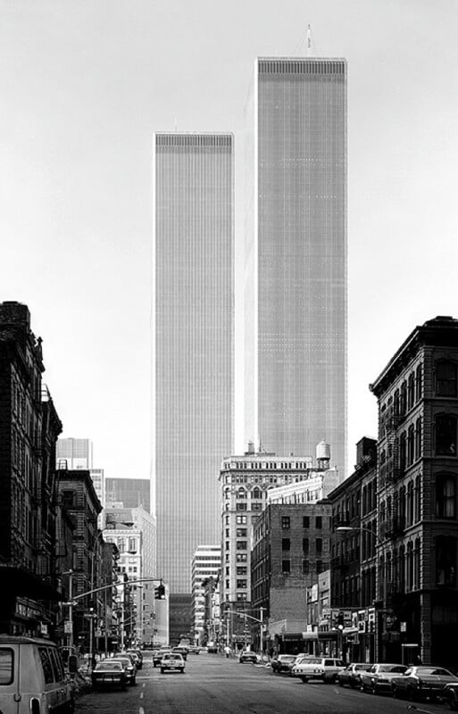 Black and white photo of the World Trade Center Twin Towers in New York City. The towers loom high above smaller buildings on either side of a broad street lined with parked cars and bustling with people. The iconic skyscrapers dominate the skyline.